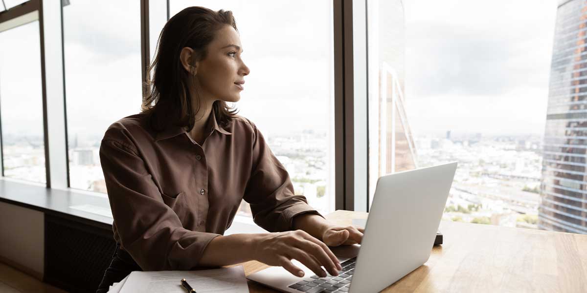woman at a desk on her laptop in her office staring out large windows at the city and pondering if seo is the right career for her or not