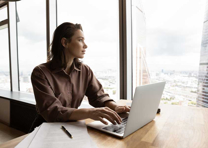 woman at a desk on her laptop in her office staring out large windows at the city and pondering if seo is the right career for her or not